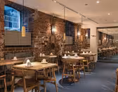 A cozy restaurant interior with exposed brick walls and warm lighting. Wooden tables and chairs are neatly arranged, each set with a folded napkin. Large windows illuminate the space, and a mirror reflects part of the dining area.