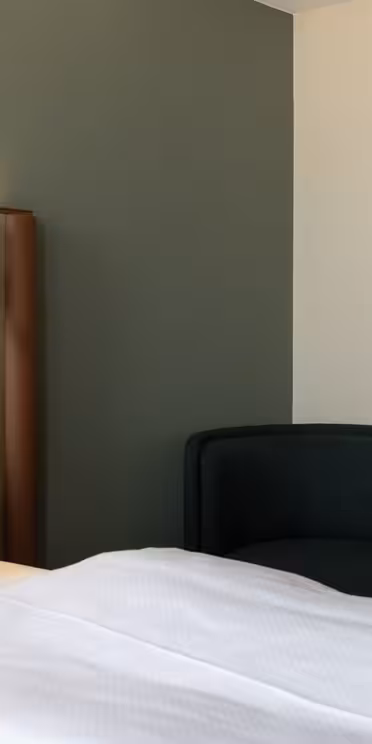 Minimalist hotel room with a bed featuring white linens, a brown padded headboard, a small round side table, a dark green chair, and a modern wall-mounted lamp against a two-tone wall.