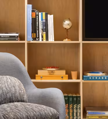 A cozy reading nook with a gray armchair in front of a wooden bookshelf. The shelves hold books, a small globe, a scrabble game box, and a decorative orange box. A television screen is partially visible on the top right.