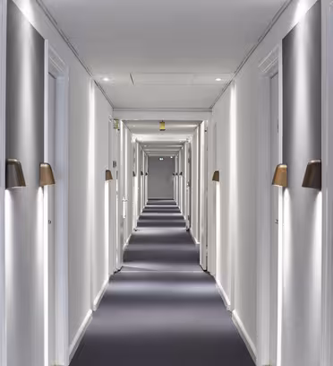 A long, well-lit hotel hallway with a dark carpet and white walls. Several doors line each side, each with wall-mounted lights above them. The hallway leads to a distant, slightly open door, creating a sense of depth.