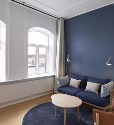 A modern living room with a blue accent wall, a blue sofa adorned with cushions, a wooden coffee table, and leather chairs. Two floor lamps are beside the sofa. Large windows offer a view of a river and buildings outside.