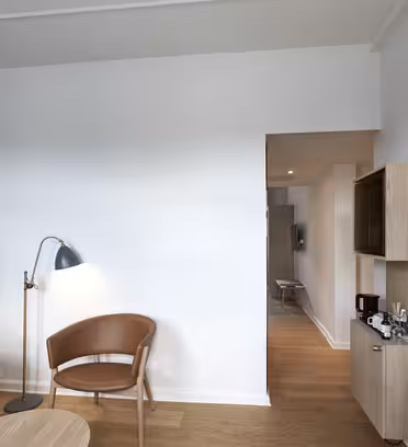 Modern hotel room with a blue accent wall on the left, featuring a brown chair and floor lamp. A wall-mounted TV and a wooden cabinet with a coffee maker are on the right. A hallway leads to another area, and the floor is light wood.