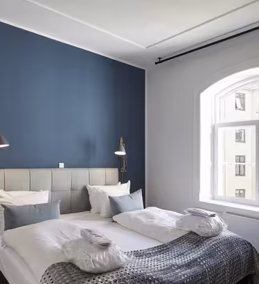 A modern bedroom with a blue accent wall, featuring a neatly made bed with white and gray bedding. There are two wall-mounted reading lamps, a small wooden side table, and a large window with a view of nearby buildings.
