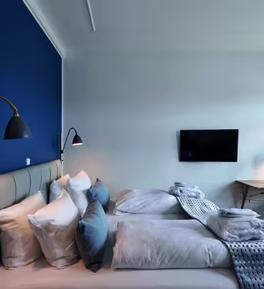 A modern hotel room featuring two neatly made single beds with a padded headboard against a blue accent wall. A flat-screen TV is mounted on a white wall. A desk with a lamp and chair is positioned by a large window with sheer curtains.
