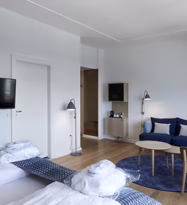 Modern hotel room with twin beds, a wall-mounted TV, and a seating area. The seating area features a blue sofa, a brown chair, and a round coffee table on a blue rug. White walls and wood floors create a clean and airy atmosphere.