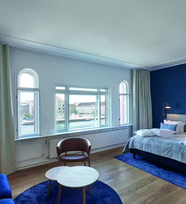A modern hotel room with a blue accent wall features a double bed, neatly made with white linens and gray blankets. Theres a blue sofa, a brown leather chair, and a small round table on a wooden floor, with large windows offering a city view.