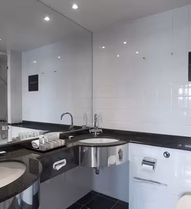 Modern bathroom with glossy white tiles, a large mirror, and black countertops. Features dual sinks, a silver faucet, toiletries, and a wall-mounted toilet. A reflection shows a glass shower. Bright lighting illuminates the space.