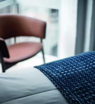 A blurred background featuring a brown chair near a window. In the foreground, there is part of a blue textured blanket draped over the edge of a light-colored bed.
