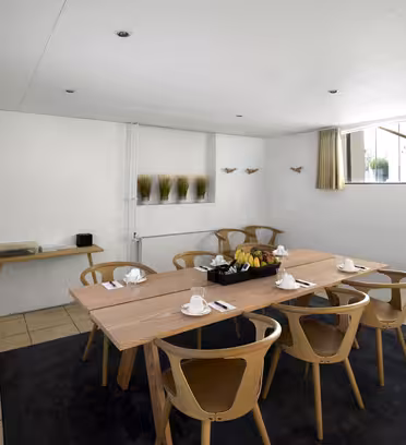 A well-lit meeting room with a wooden table, surrounded by eight chairs. The table is set with cups, saucers, and a fruit basket. A wall-mounted flat-screen TV is on the left, and there are tall windows with beige curtains above.