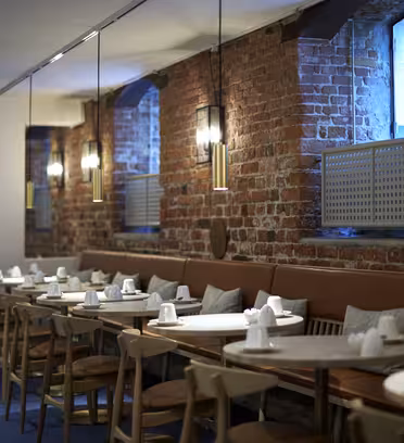 Cozy restaurant interior with exposed brick walls and long leather seating. Several tables are set with napkins and cutlery. Hanging pendant lights provide warm lighting, and large windows let in natural light.