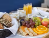 A breakfast tray featuring a variety of fresh breads in a basket, a plate with sliced cantaloupe, oranges, apples, grapes, and plums, three glasses of yogurt or parfaits, and two glasses of orange juice. A blue cushion is in the background.