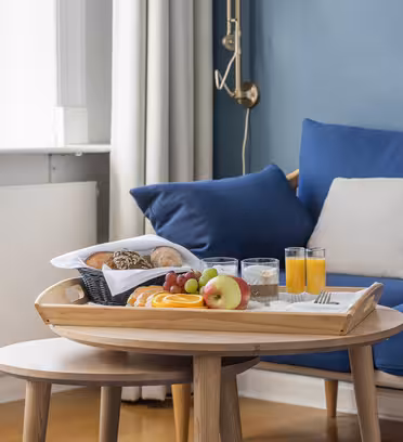 A wooden tray on a round table holds a breakfast selection including sliced fruit, bread rolls, and two glasses of juice. A cozy blue sofa with beige pillows is in the background, near a window with cream curtains.